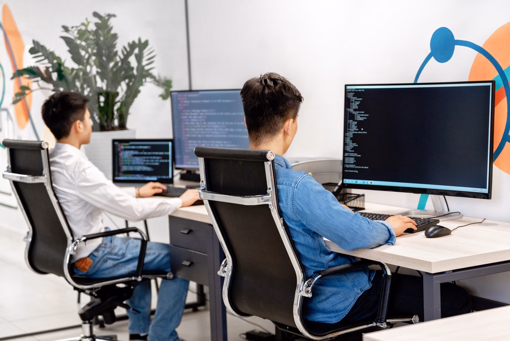 Two people working at separate desks on computers with code displayed on their screens.