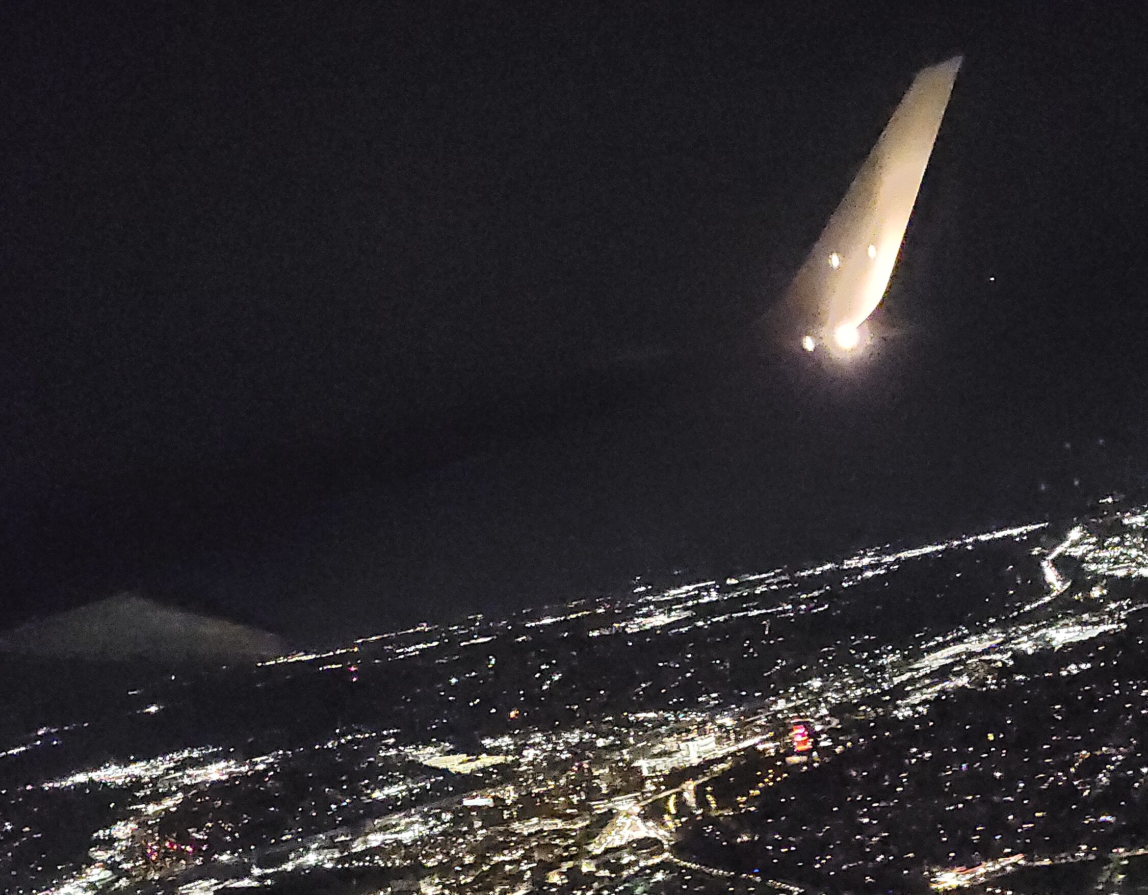 This is a decorative image. Picture at night with complete darkness. There is a light illuminating a plane wing. Below the plane wing is a lit up city that also adds light to the image.